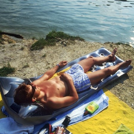 A sunburned man lies shirtless on a pool float by a lake, tanning under direct sunlight.