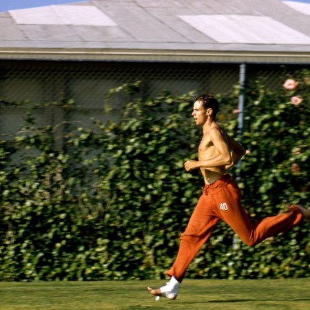 Shirtless male runner in red track pants sprinting on grass with gauze around his feet.